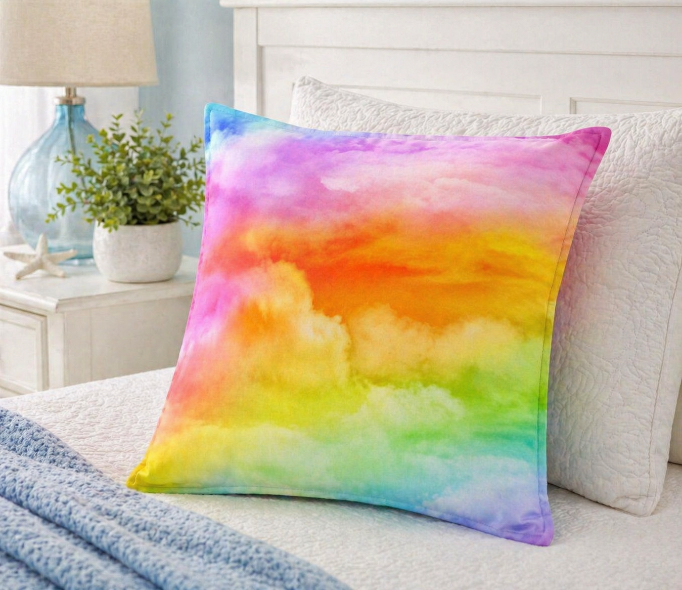 A square throw pillow with a vibrant rainbow watercolor cloud pattern, styled on a bed with white bedding and a blue blanket next to a bedside table with a lamp and plant.