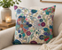 A square throw pillow with a vibrant, multicolored floral and paisley pattern on a cream background, styled on a beige sofa next to a lit candle and a potted plant.