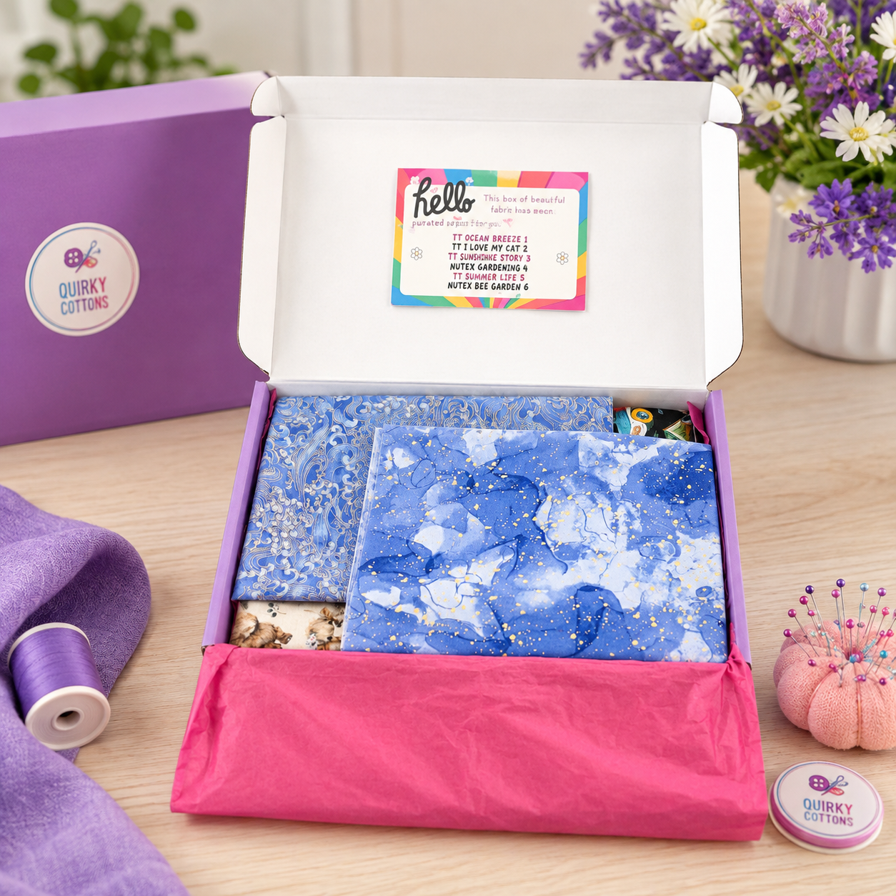 An open purple Quirkly Cutting gift box on a wooden table, showing blue patterned fabric inside with a pink tissue paper lining. A purple box lid and spool of purple thread sit to the left, and a pink pumpkin-shaped pin cushion with daisies and a small Quirkly Cutting pin case sit to the right.