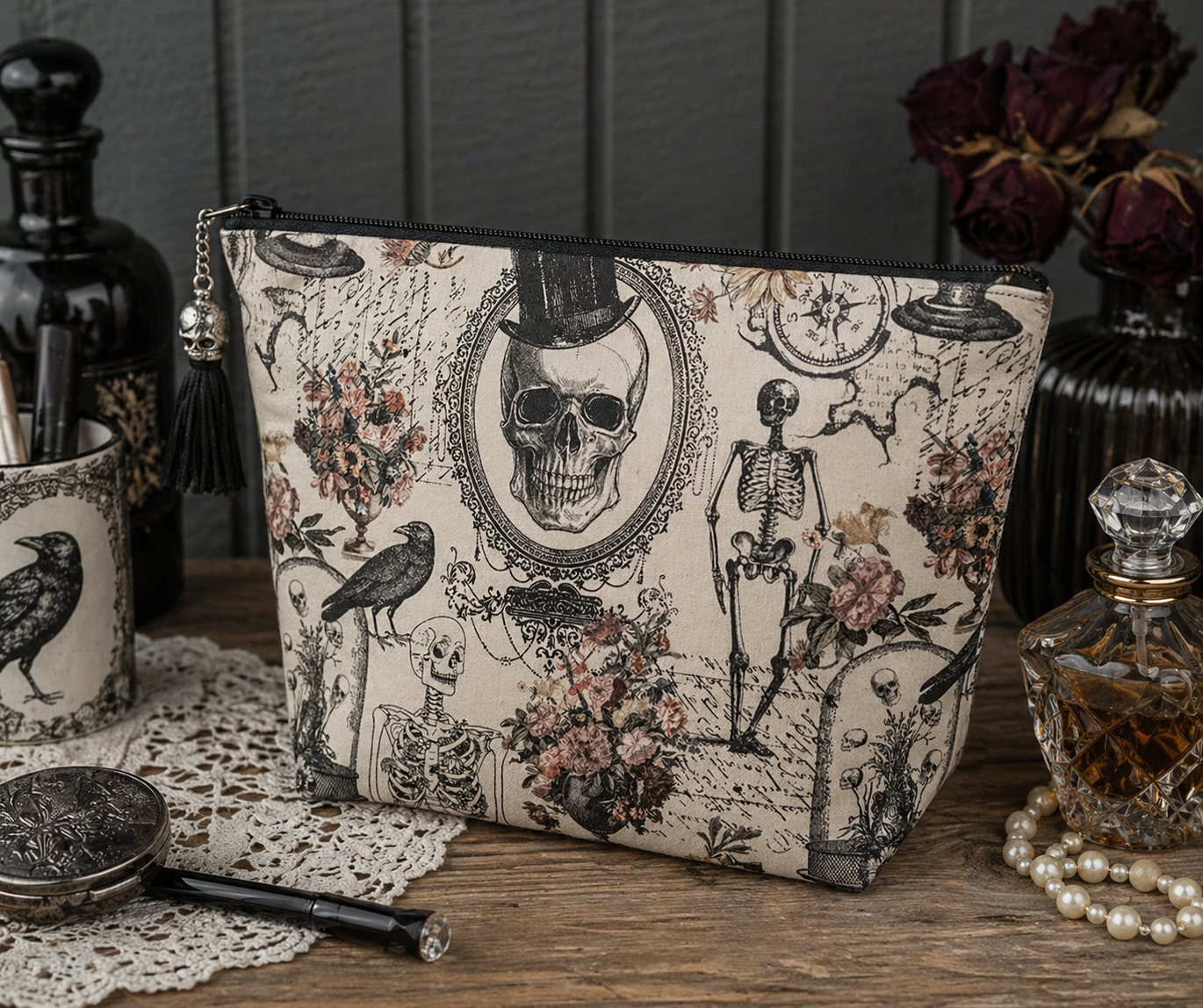 A rectangular cosmetic pouch with a gothic skeleton and skull print, styled on a wooden table with vintage props including a perfume bottle, a mirror, and dried flowers.